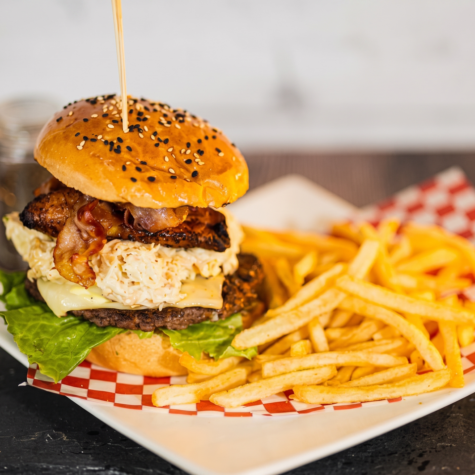 A gourmet burger with bacon, cheese, and mac and cheese, served with fries on a checkered paper.