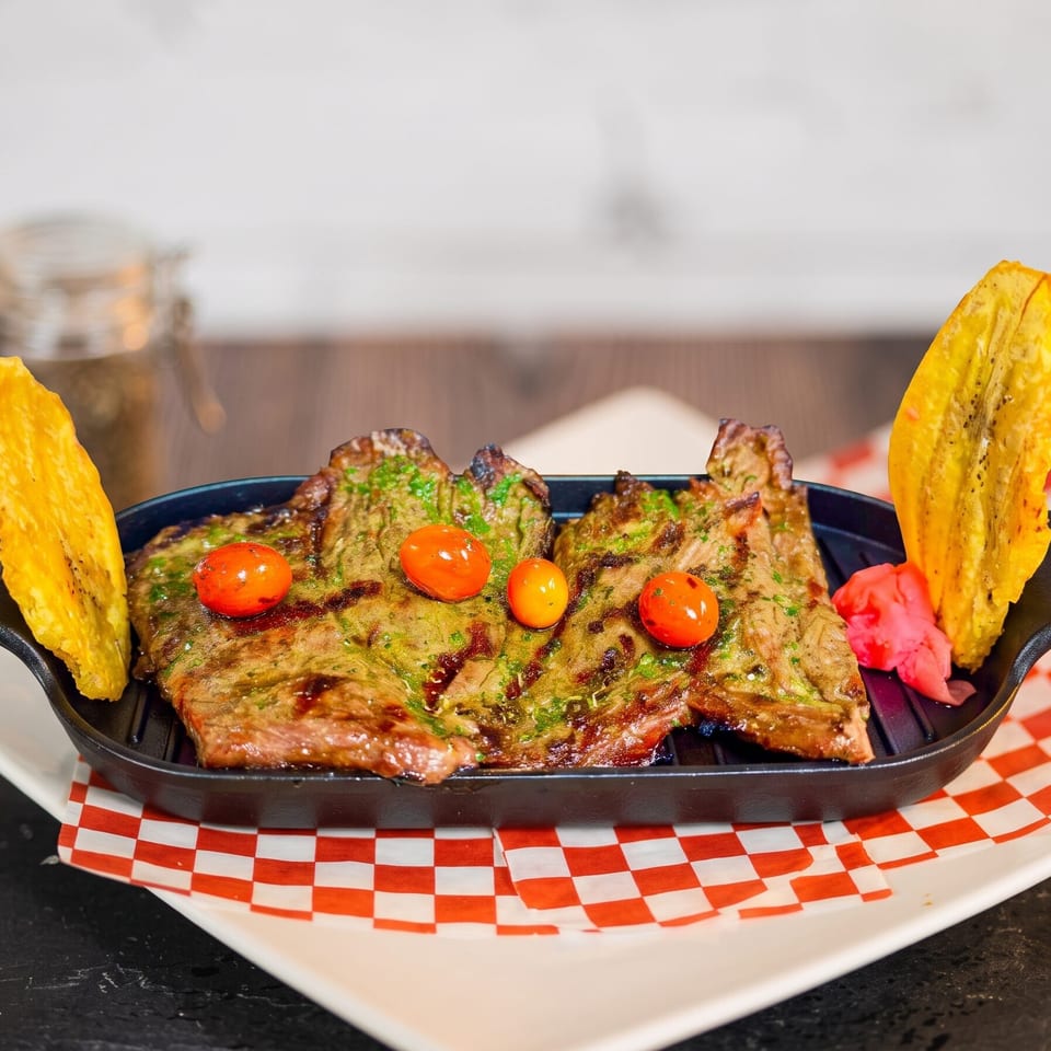 Grilled steak with cherry tomatoes and plantain slices in a cast iron skillet on a checkered paper.