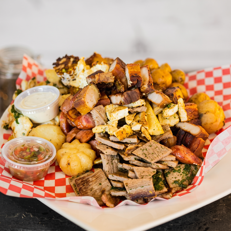 A large platter of grilled meats, sausages, potatoes, and sauces on a red and white checkered paper.